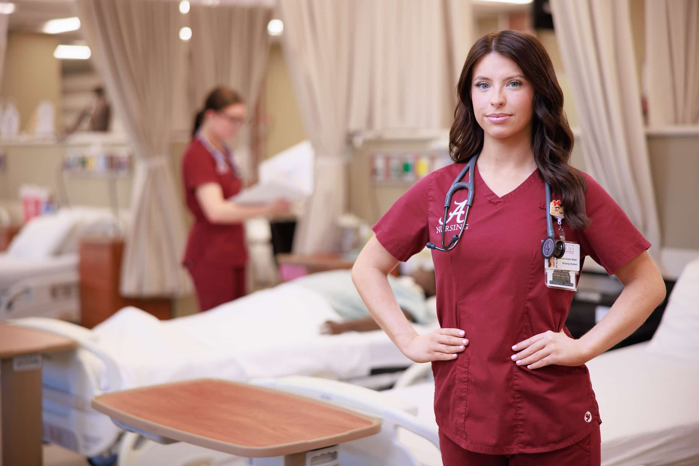 Nursing student posing in simulation lab with fellow student examining a nursing mannequin in the background