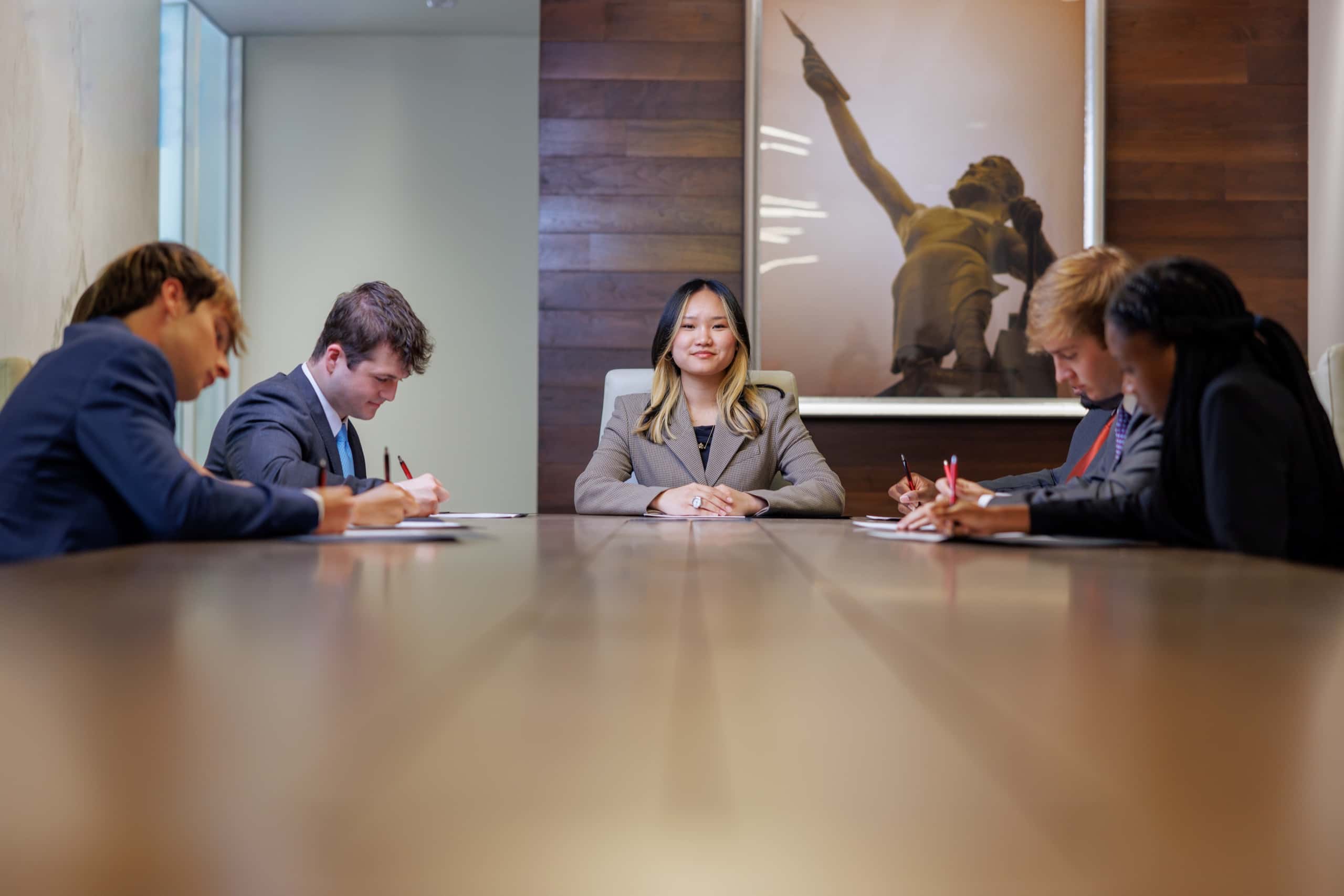 Five students in business attire seated around a conference table, one smiling toward the camera as the others take notes.
