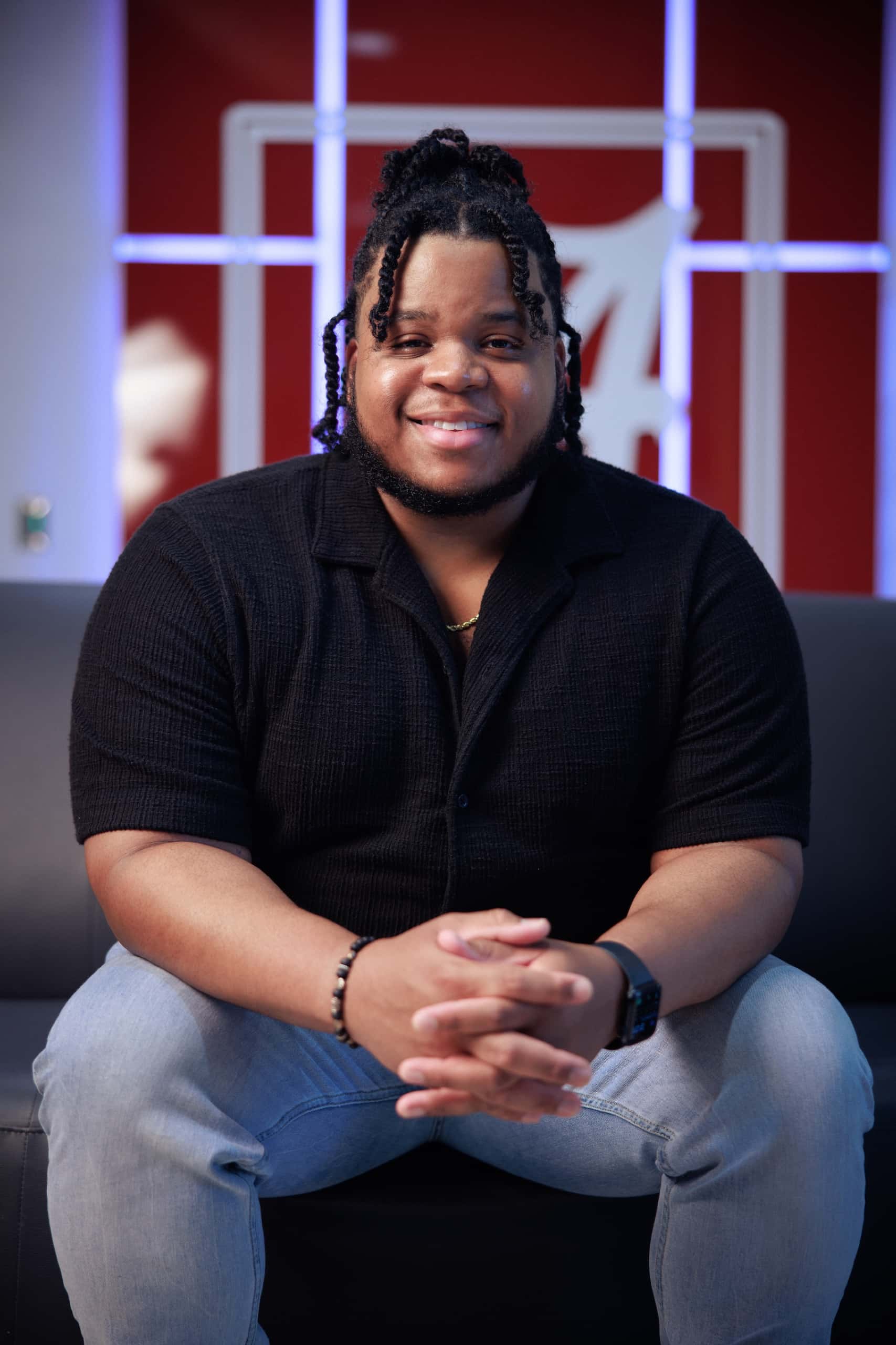 A smiling student in a black shirt sits with hands clasped in front of a crimson wall with the script A logo.