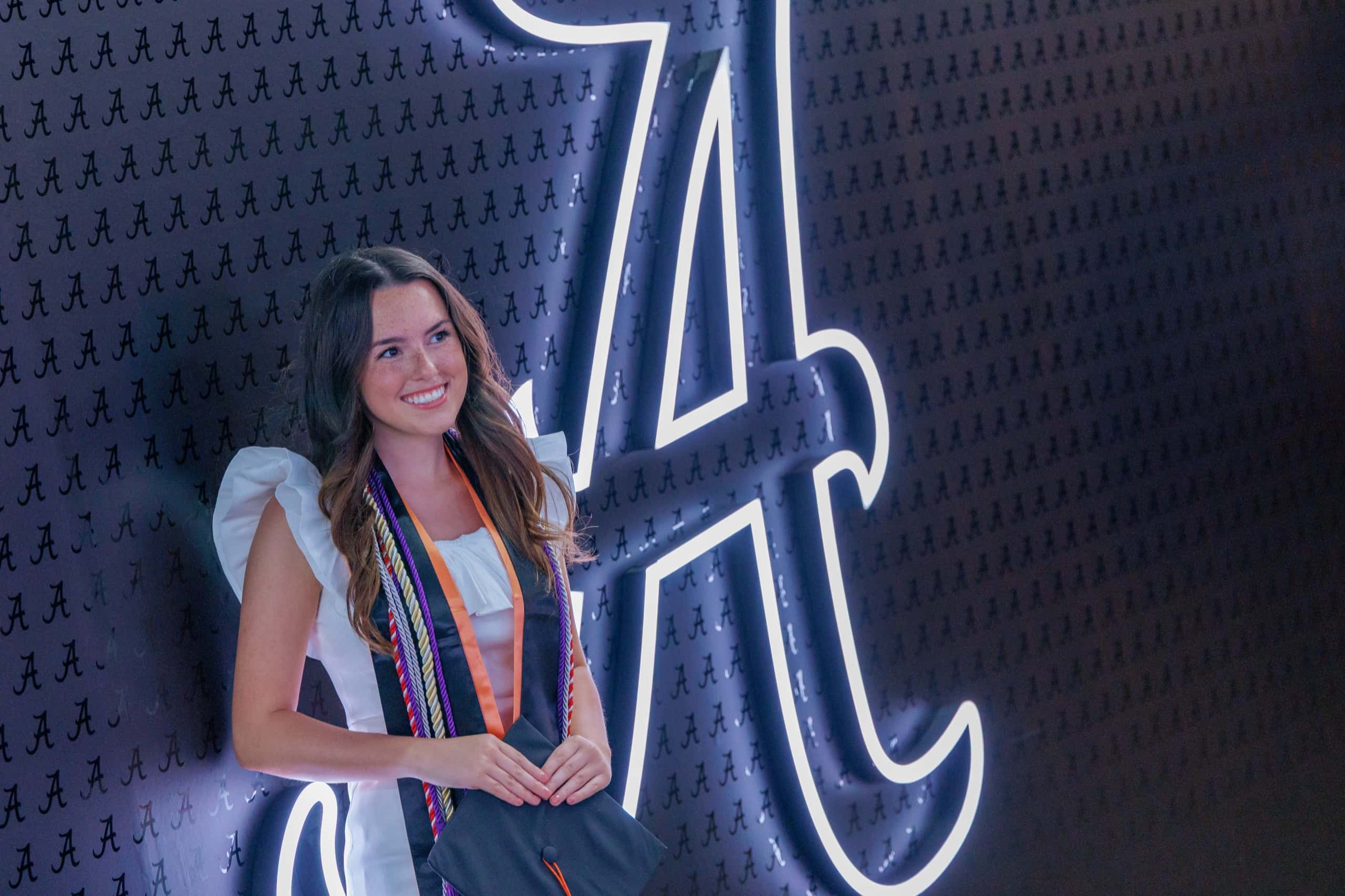 A graduate in cap and gown with honor cords smiles in front of a glowing script A wall.