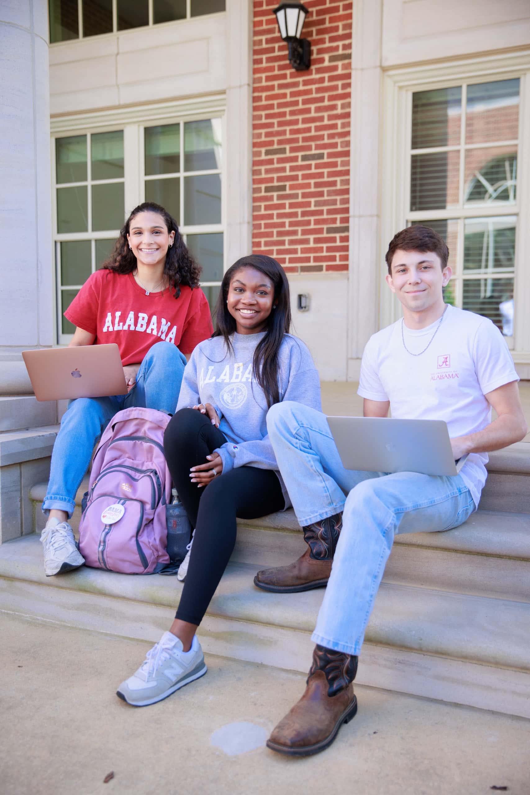 Three students in Alabama apparel sit on building steps with laptops, smiling toward the camera.