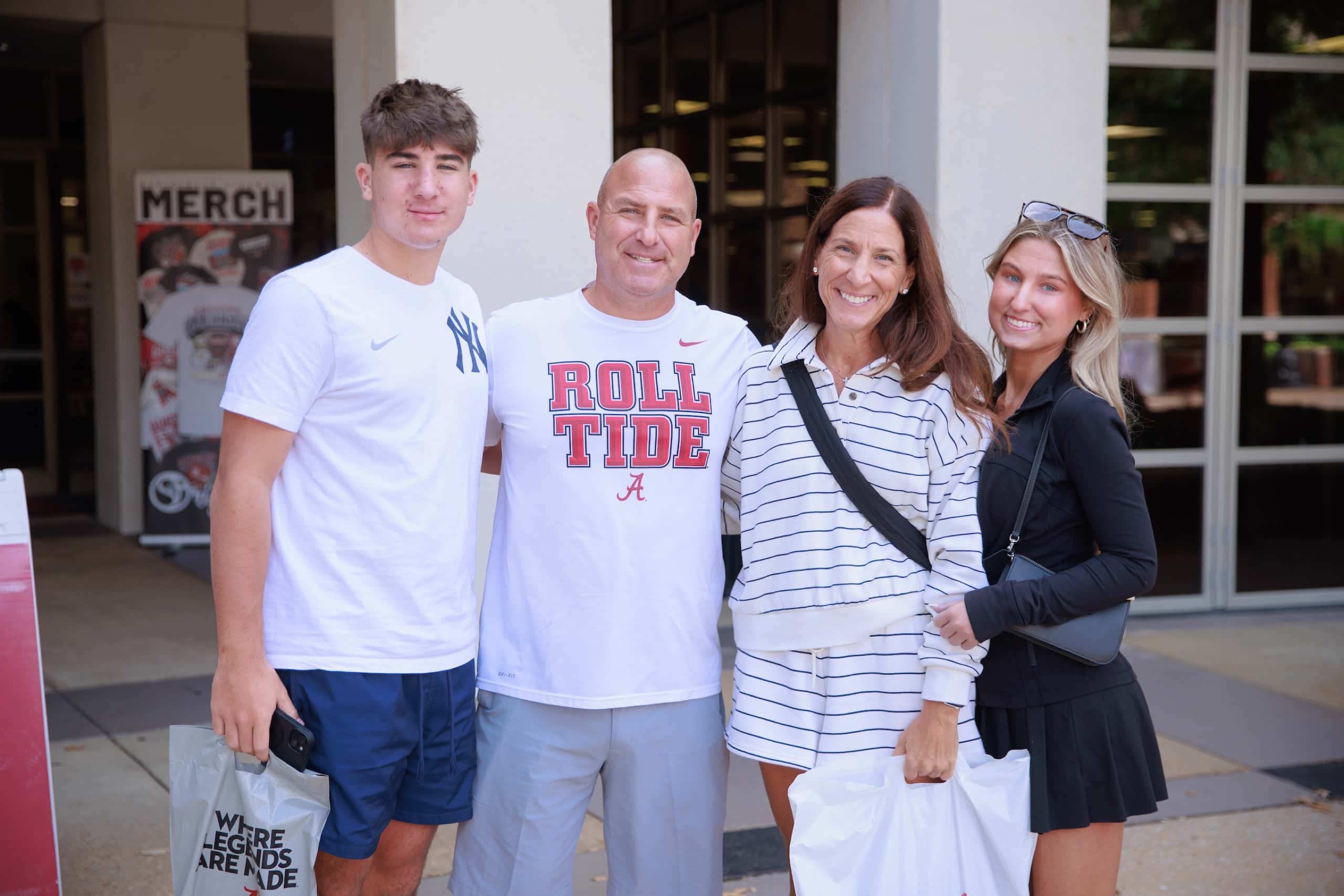 A family of four smiles together outside a campus store, one in a Roll Tide shirt, holding shopping bags.