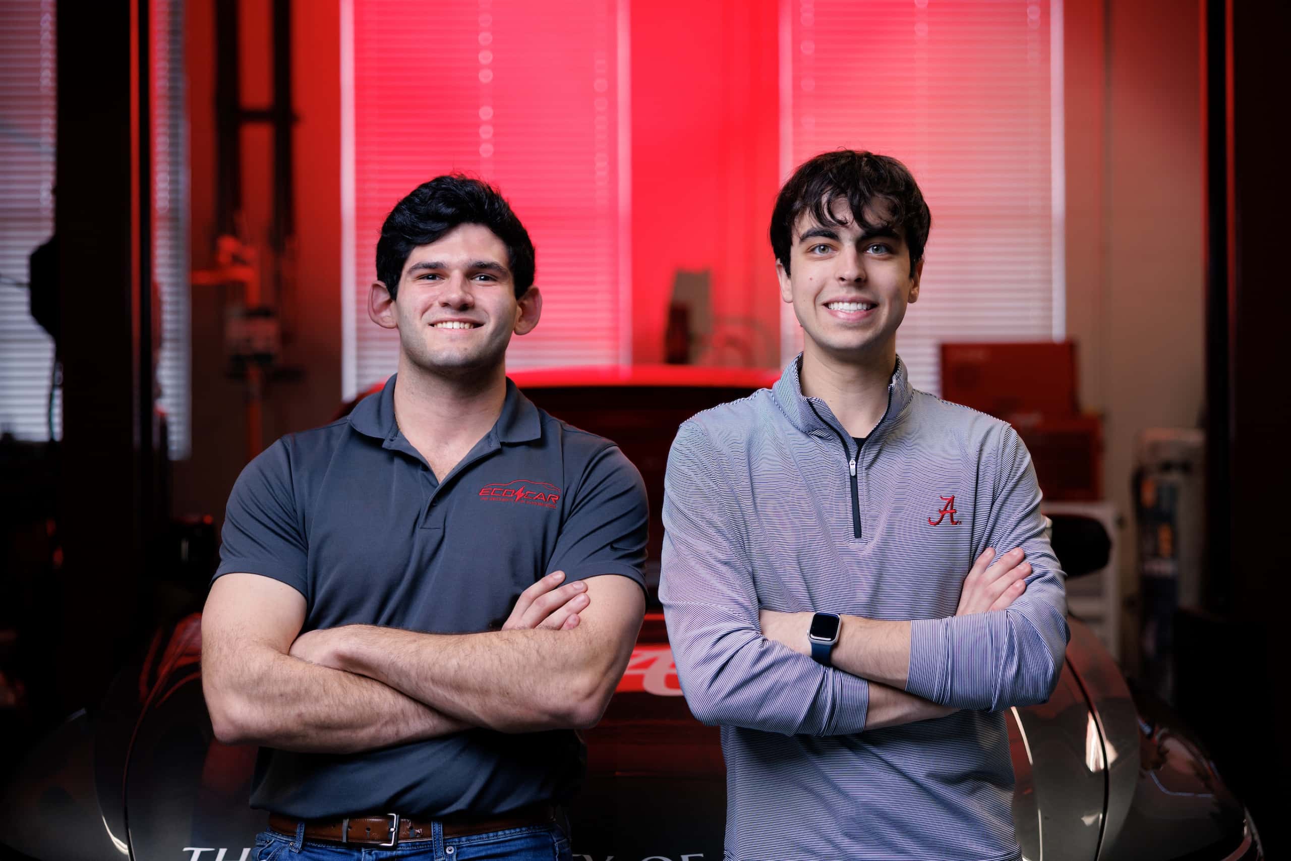 Two students stand with arms crossed in front of an EcoCAR competition vehicle in a campus engineering lab.c
