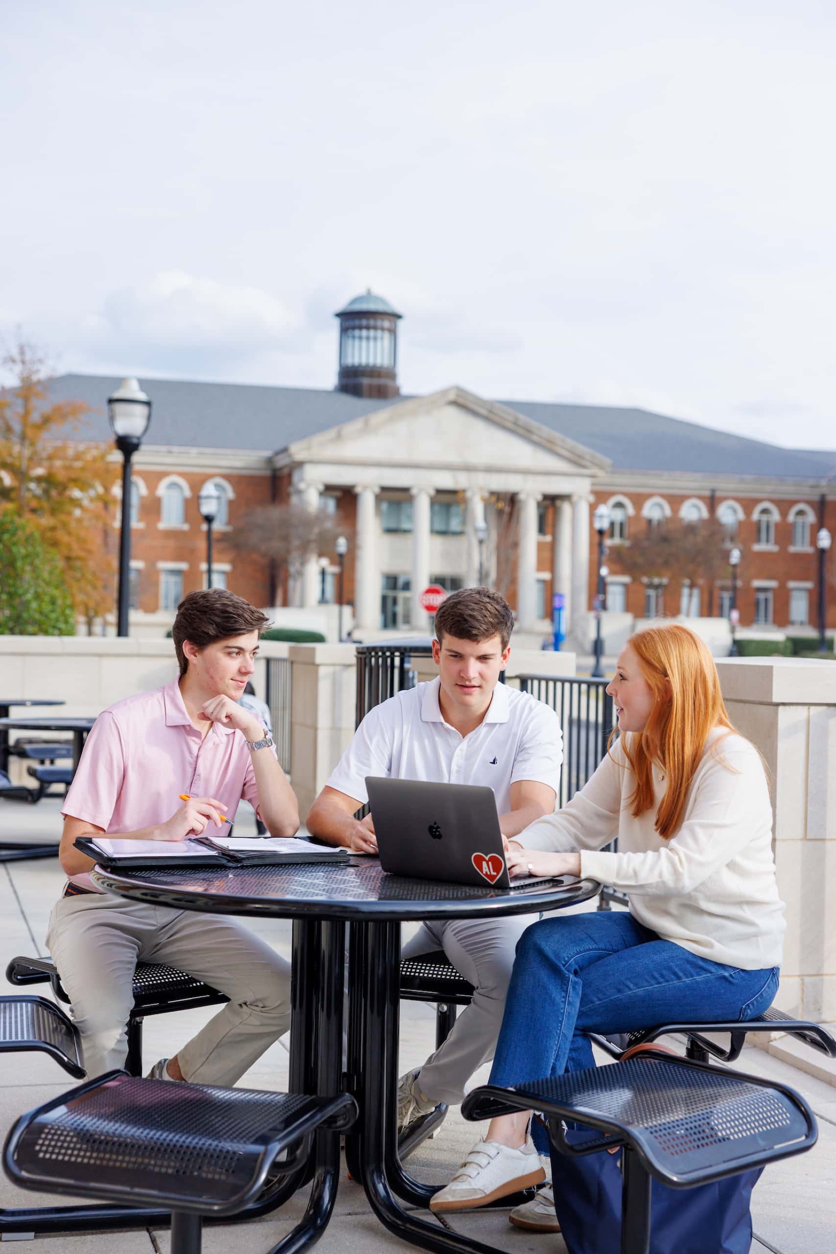 Three students talk around an outdoor table with a laptop, a historic brick campus building behind them.