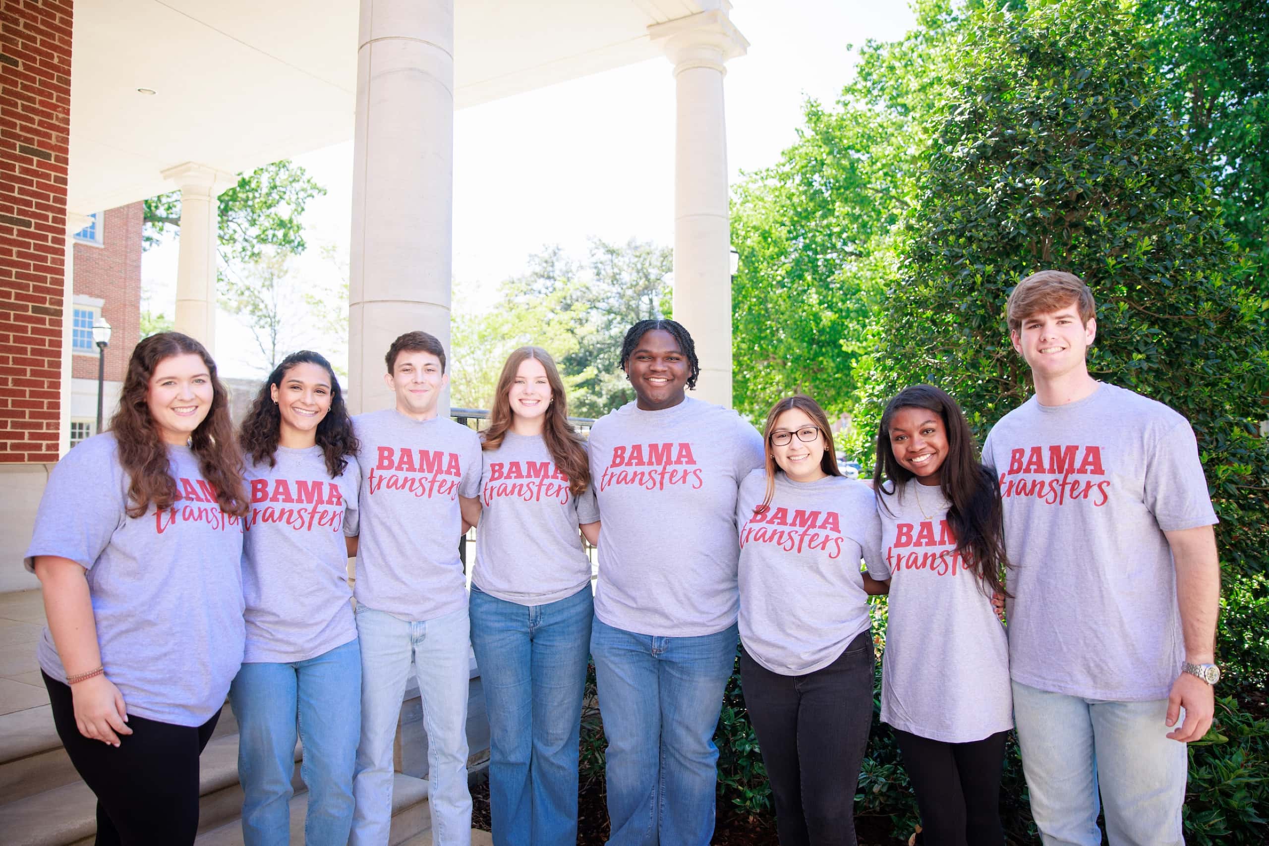 Eight students in gray "Bama Transfers" T-shirts stand together and smile beneath columns outside a campus building.